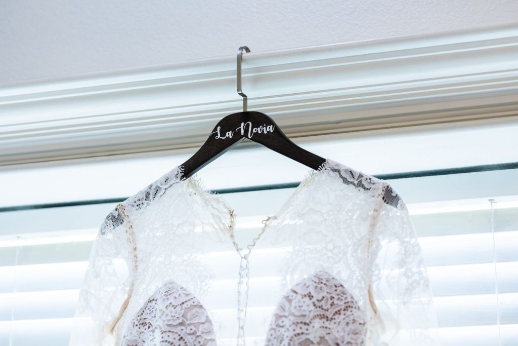 Elegant lace bridal gown with intricate details hanging on a personalized wooden hanger indoors.