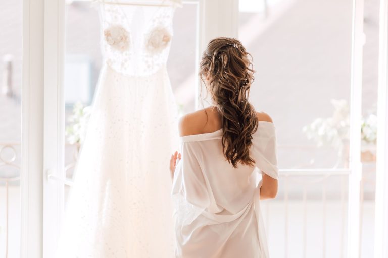A bride stands with her back turned, gazing at her wedding dress in a serene indoor setting.