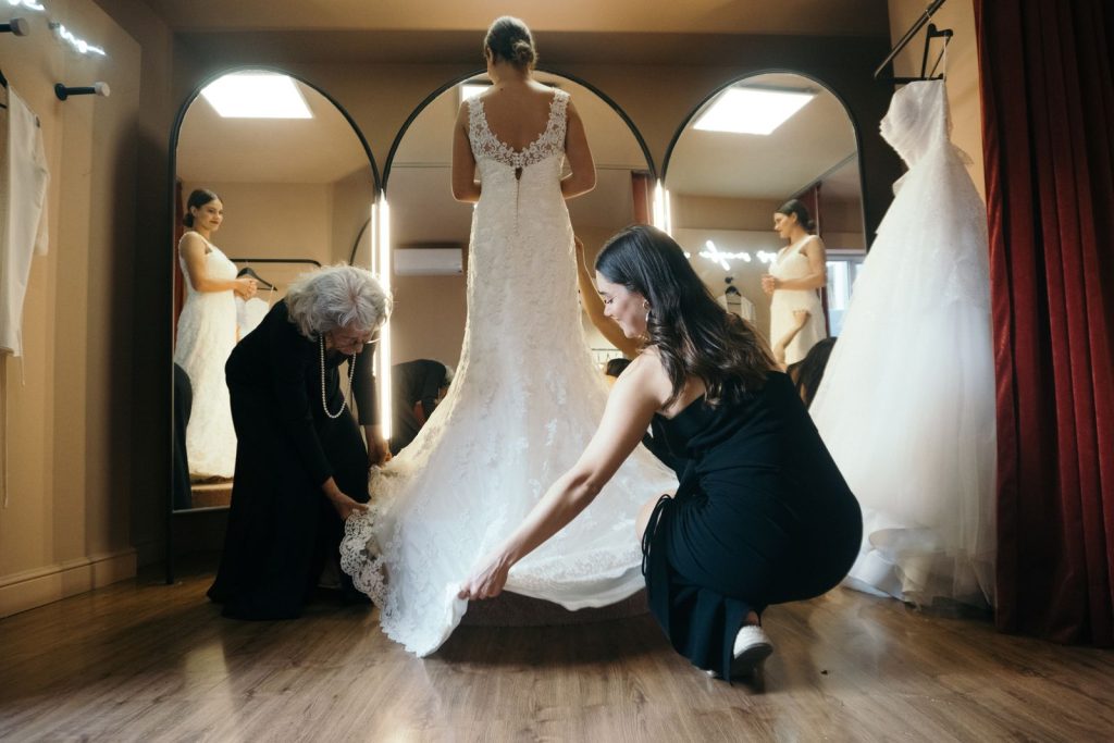 A bride stands in a fitting room as two women assist with her wedding dress adjustments.