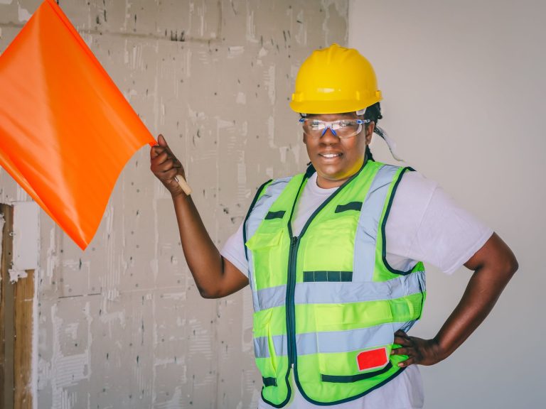A black female construction worker holds a flag, wearing safety gear, hardhat and vest.