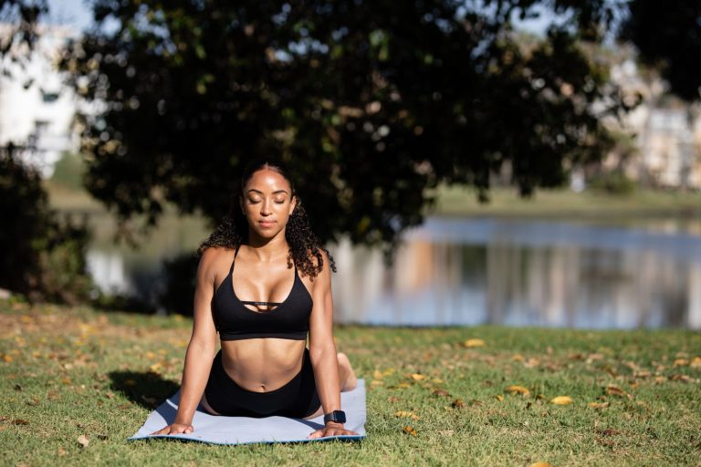 A woman practicing yoga on a mat outdoors by the lake on a sunny day, embracing wellness.