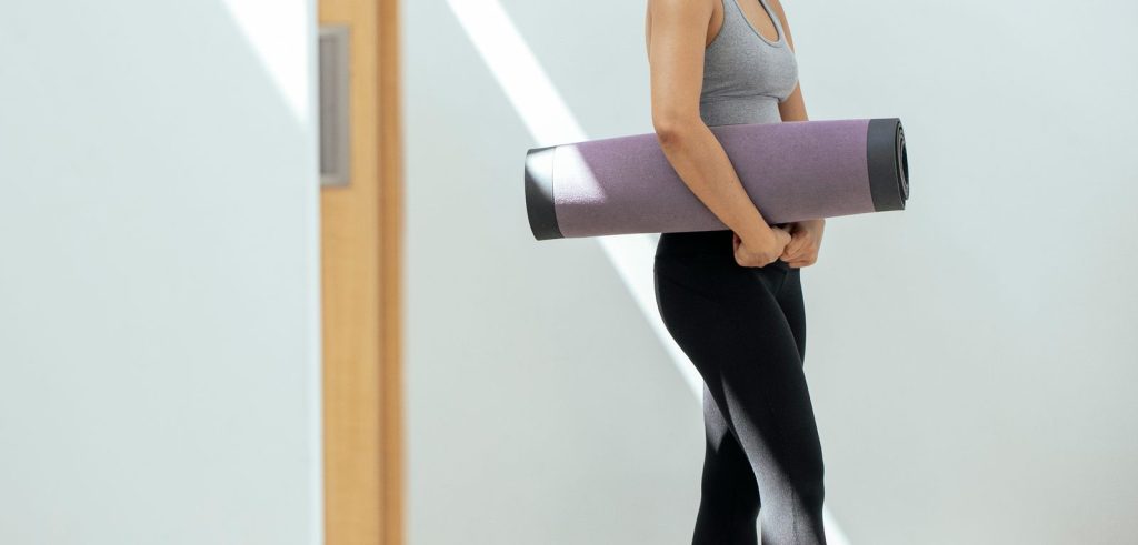 Side view of faceless young fit female in sportswear standing near white wall with rolled mat in hand after finishing yoga training in light studio
